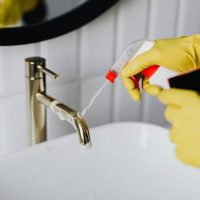 From above anonymous housekeeper in yellow rubber gloves spraying cleaner to chrome water faucet in bathroom